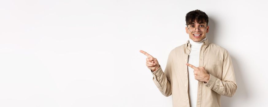Cheerful handsome man in glasses smiling, pointing fingers left at logo, showing advertisement with excited face, standing on white background.