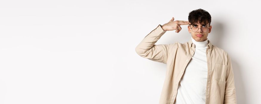 Annoyed young man making finger gun gesture on head, shooting himself from boredom or annoyance, tired of something stupid, standing on white background.