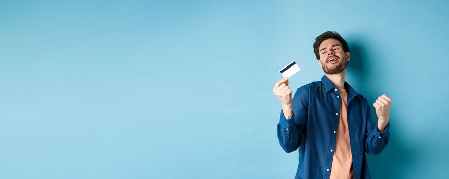 Happy and satisfied young man dancing with plastic credit card, fist pump and smile, standing on blue background.