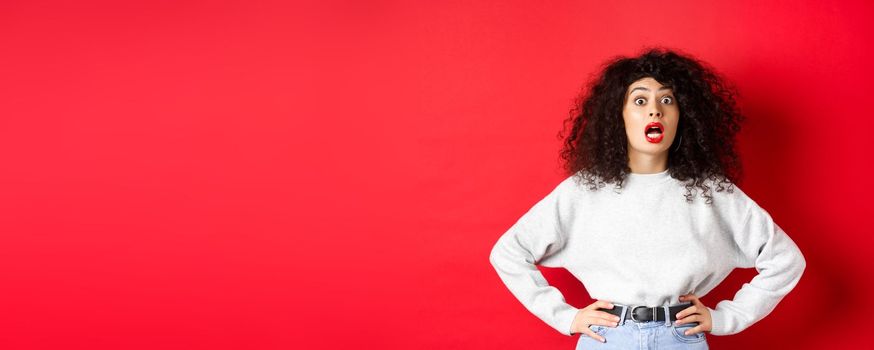 Shocked italian woman with curly hair, gasping and staring at camera astonished, open mouth, standing in white sweatshirt on red background.