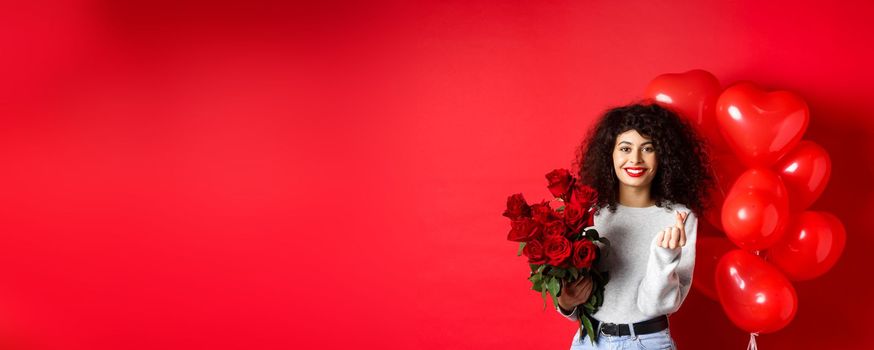 Holidays and celebration. Beautiful girlfriend receive flowers on anniversary, showing finger heart and standing near party balloons, red background.