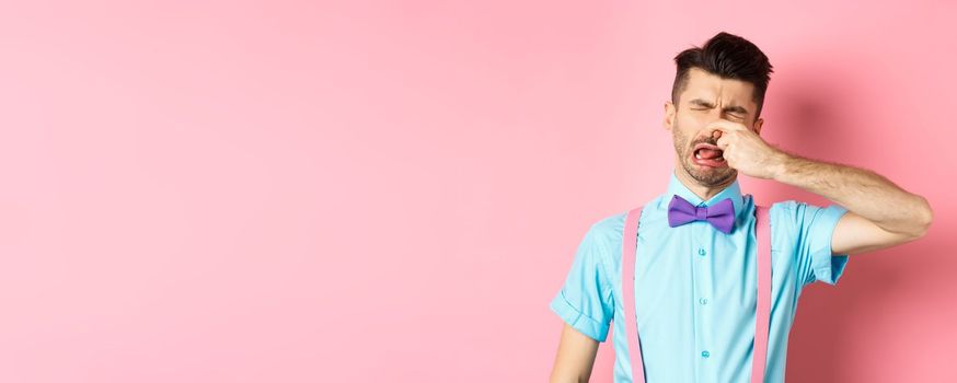 Image of man crying from disgusting smell, shut his nose from awful stink, standing in bow-tie and suspenders on pink background.