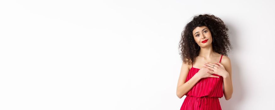 Touched young woman with curly hair, wearing red dress, holding hands on heart and smiling grateful, say thank you, standing over white background.