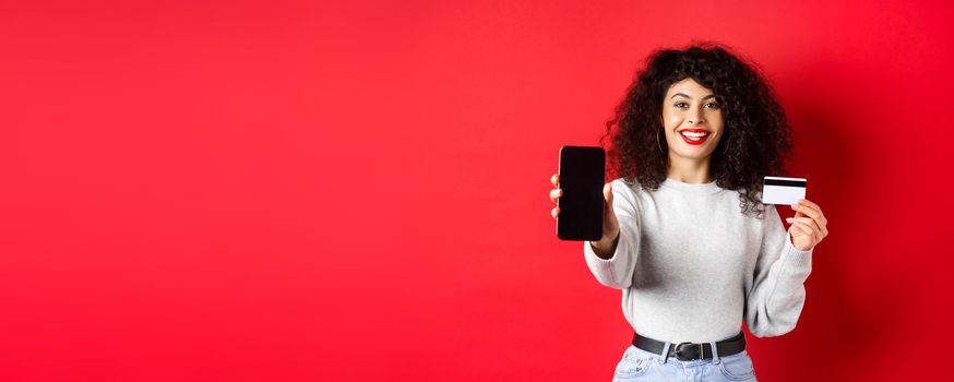 Happy good-looking girl showing plastic credit card and empty smartphone screen, demonstrate account or application, standing on red background.