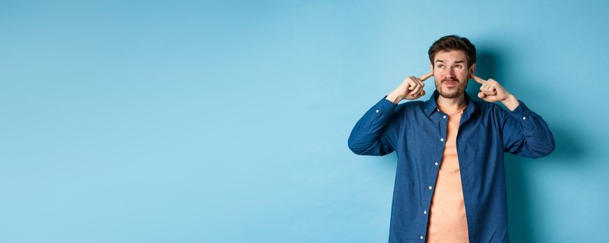 Annoyed young man block sound, shut ears with fingers and grimacing, disturbed by loud music, standing on blue background.