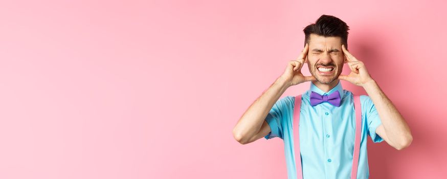 Image of man with headache touching head temples, grimacing from painful migraine, clench teeth, standing sick on pink background.