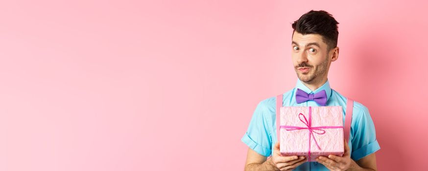 Holidays and celebration concept. Funny guy with moustache showing cute gift box wrapped in festive paper, looking at camera, presenting you, standing on pink background.