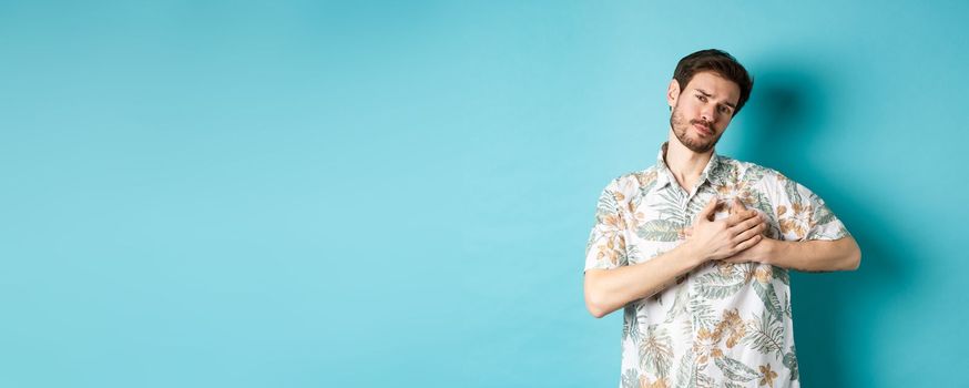 Passionate and romantic guy in hawaiian shirt, holding hands on heart and looking with sympathy, being in love, standing on blue background.