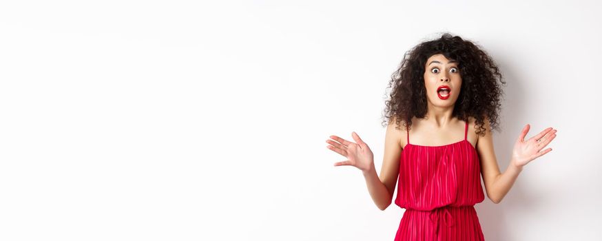 Excited woman with curly hair, talking with amazed face, explain big news, shaking hands and look astonished at camera, white background.
