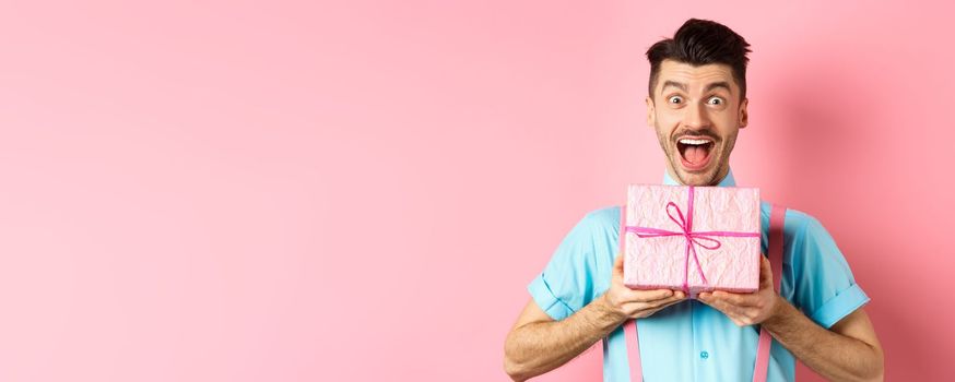 Holidays and celebration concept. Excited and surprised guy celebrating birthday, receiving gift and cheering, smiling happy at camera, standing over pink background.