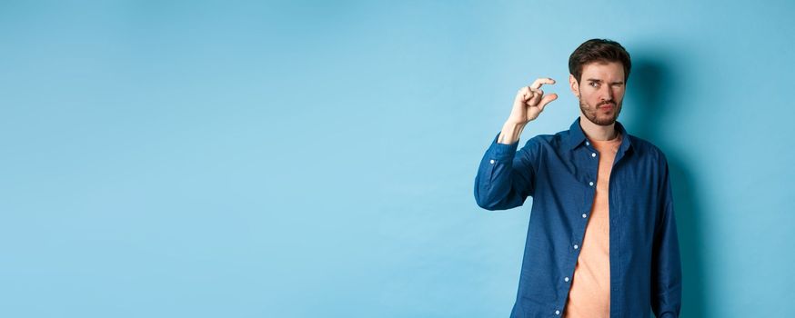 Image of young caucasian man showing small size and squinting to see something tiny, standing on blue background.