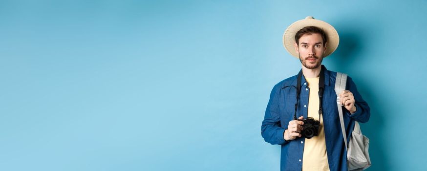 Handsome young tourist in summer hat, backpacking and travelling, holding photo camera, standing on blue background.