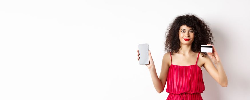 Online shopping. Fashionable woman with curly hair, wearing red dress, showing smartphone screen and plastic credit card, smiling at camera, white background.