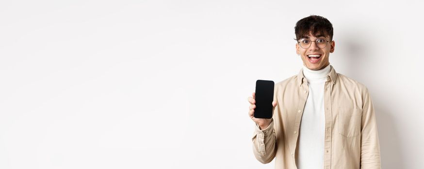 Technology and online shopping concept. Cheerful handsome guy in glasses showing empty smartphone screen and smiling, showing application, standing on white background.