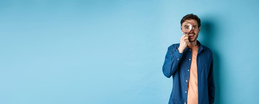 Cheerful young man found something good, smiling and looking through magnifying glass, standing on blue background.