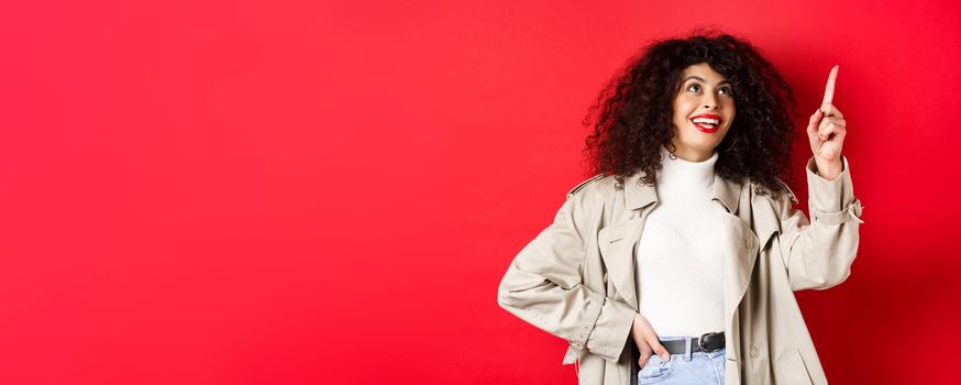 Excited woman with curly hair, wearing trench coat, pointing at upper right corner and smiling amazed, standing against red background.