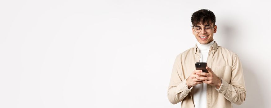 Real people. Natural young man reading message on mobile phone, smiling and looking at smartphone screen, standing on white background.