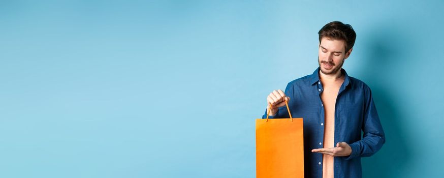 Young smiling guy showing orange shopping bag, demonstrating gift, standing on blue background.