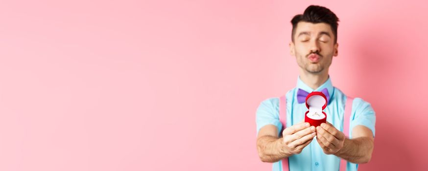 Valentines day. Silly boyfriend in bow-tie pucker lips, close eyes and waiting for kiss after making proposal, showing golden engagement ring in small red box, standing on pink background.