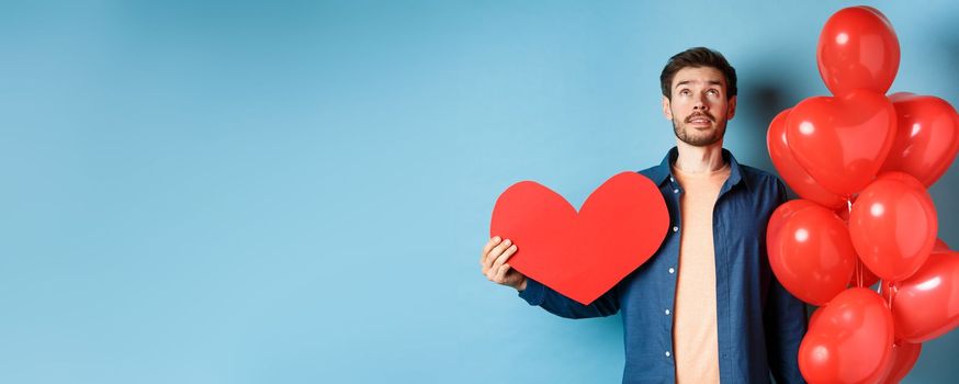 Valentines day and love concept. Man looking up with dreamy face, holding romantic gift balloons and red heart cutout, standing over blue background.