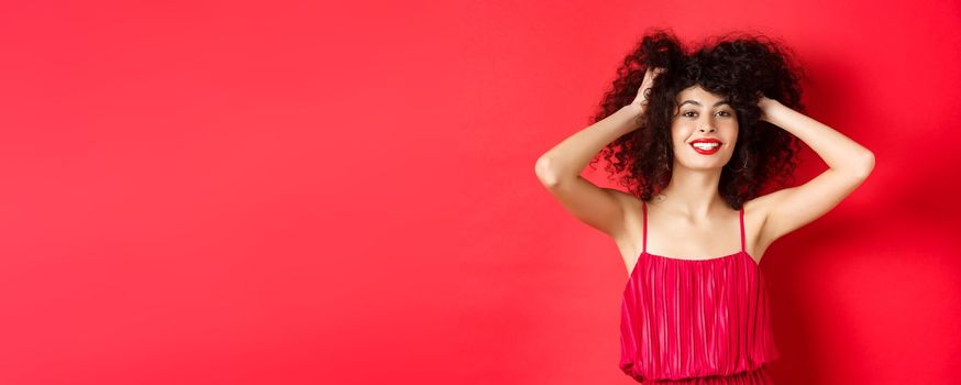 Carefree lady in red dress, touching her curly hair and smiling happy, standing on studio background. Copy space