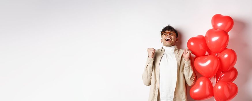 Valentines day. Happy modern man celebrating, screaming from joy and happiness, having date with lover, being in love, standing near heart balloons on white background.