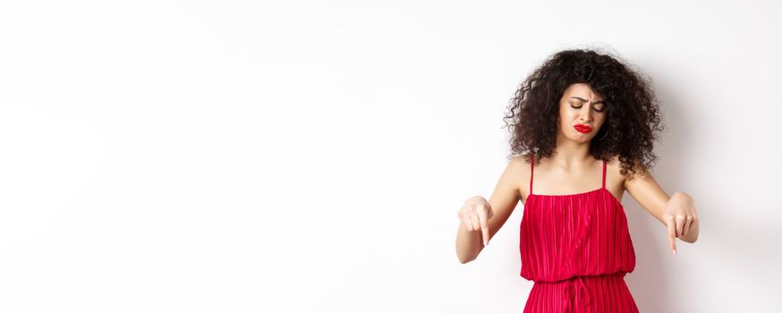 Sad and disappointed curly-haired woman in red dress, frowning and looking down jealous, pointing at promo, standing over white background.