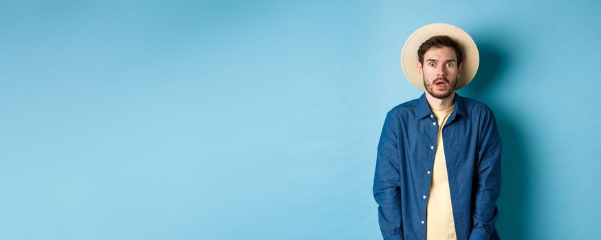 Young man tourist in summer hat drop jaw, staring at something shocking, standing on blue background.