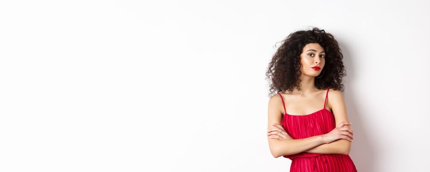 Sassy woman with curly hairstyle, wearing red dress and makeup, cross arms on chest, standing over white background.