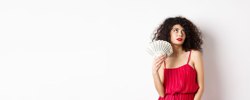 Elegant beautiful woman in red dress, waving with dollars and looking fabulous, standing on white background. Copy space