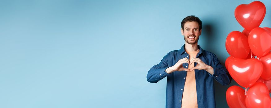 Happy valentines day. Boyfriend in love showing heart gesture to lover and smiling, standing near red balloons on blue background.