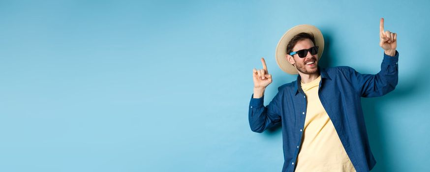 Funny tourist dancing on vacation, pointing fingers up, wearing summer hat and sunglasses, blue background.