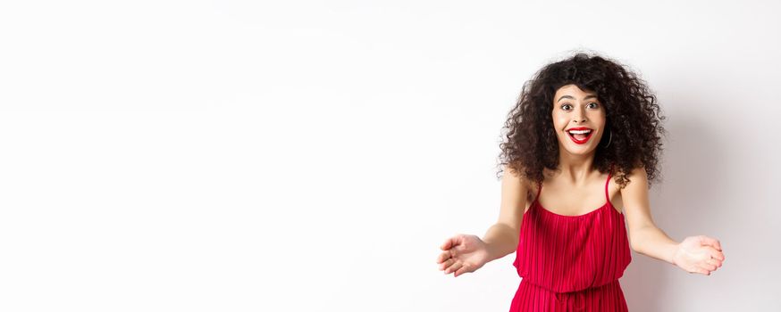 Portrait of happy caucasian woman in red dress and makeup, stretch out hands to beckon someone, inviting for hug, receiving surprise gift, standing on white background.