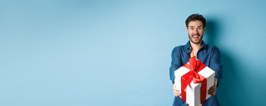 Valentines day. Excited handsome man giving gift box and wishing happy holiday. Romantic guy extending hands with present, standing againt blue background.