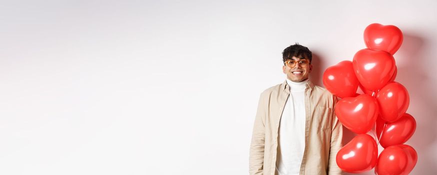 Valentines day. Excited young man smiling, looking hopeful, standing near big red hearts balloons, waiting for true love on lovers date, white background.