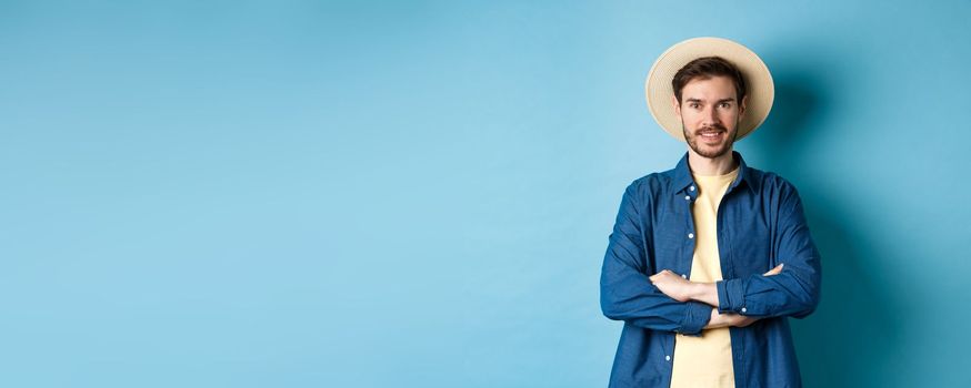 Portrait of cheerful guy going on vacation in straw summer hat, cross arms on chest and smiling, looking confident, standing on blue background.