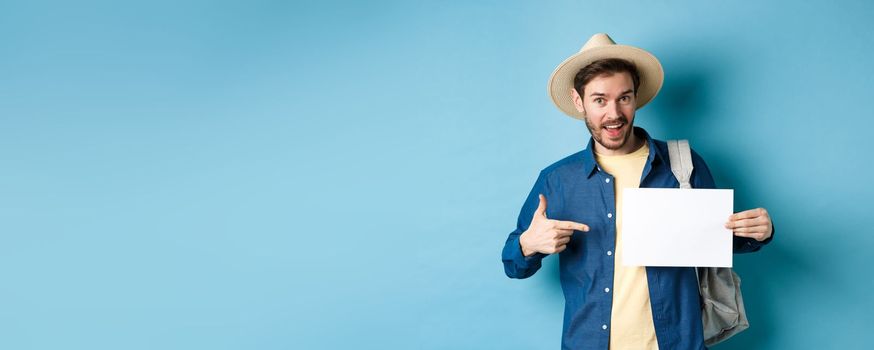 Handsome tourist smiling and pointing at empty piece of paper, wearing straw hat and backpack. Concept of summer vacation.