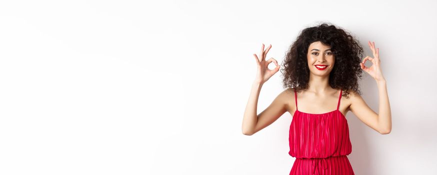 Beautiful woman in red dress, smiling and showing okay gestures, like something good, praising good job, standing over white background.