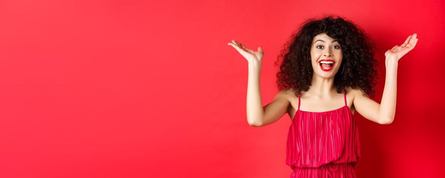 Excited beautiful woman raising hands up and scream with happiness and joy, applause with rejoice, standing on red background.