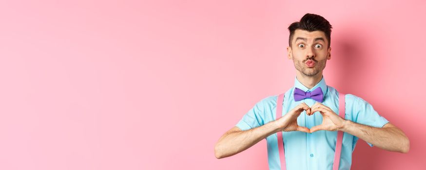 Happy Valentines day. Funny young man waiting for lovers kiss, pucker lips and show heart sign, I love you gesture, express feelings on date, pink background.