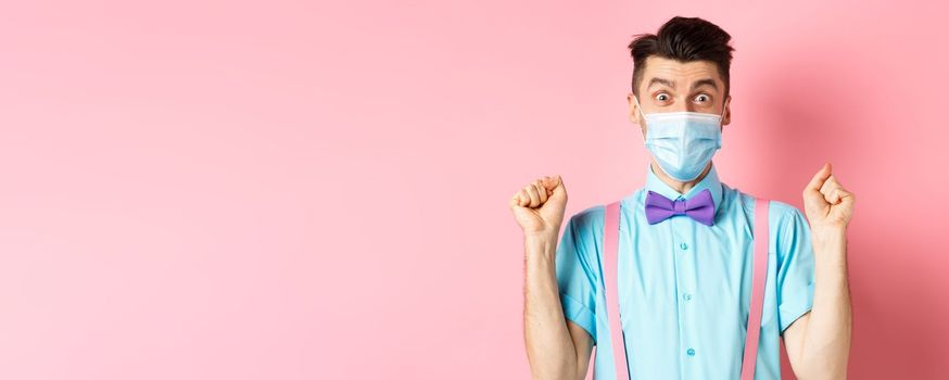 Covid, pandemic and quarantine concept. Cheerful young man in face mask winning prize, staring at camera amazed, raising hands up with hopeful expression, standing on pink background.