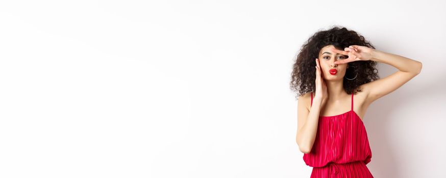 Beauty and fashion. Stylish curly-haired woman in red dress showing v-sign on eye and pucker lips, kissing you, standing on white background.