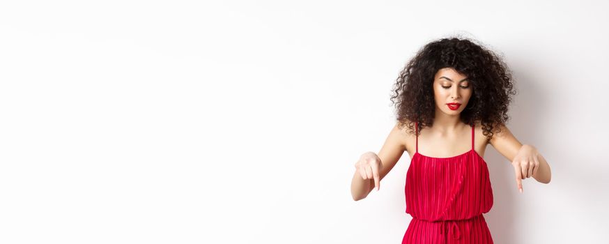 Intrigued beautiful woman with red lips and dress, pointing and looking down at banner, reading logo, standing over white background.