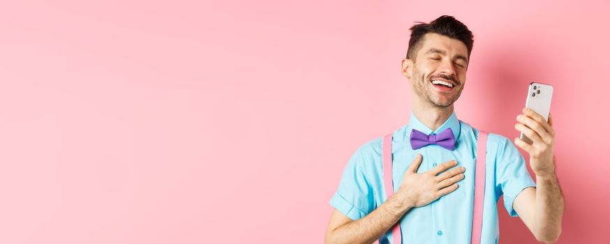 Technology concept. Happy caucasian man video chat and laugh at smartphone, standing over pink background.