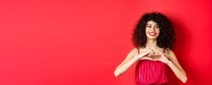 Valentines day. Romantic girl with curly hairstyle in evening dress, smiling and showing heart sign, say I love you on lovers holiday, standing over red background.