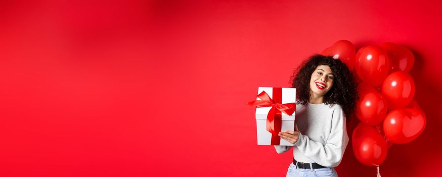 Holidays and celebration. Happy birthday girl holding gift and posing near party helium balloons, smiling excited at camera, red background.