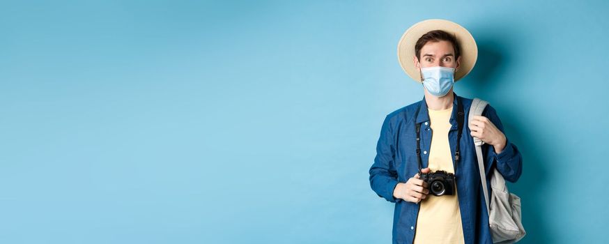 Covid-19 and travelling concept. Young guy tourist in medical mask and summer hat travel abroad during coronavirus pandemic, taking pictures on vacation, blue background.