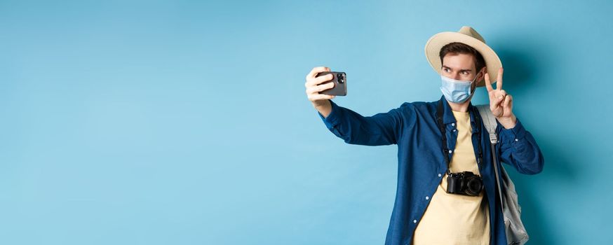 Covid-19, pandemic and travel concept. Happy and positive guy in tourist hat taking selfie and showing peace sign, posing near sightseeing, blue background.