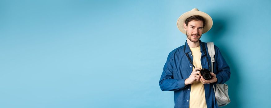 Handsome guy going on summer vacation, backpacking on holiday. Tourist with straw hat and camera smiling happy, standing on blue background.