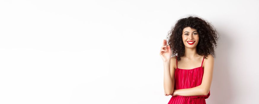 Beauty and fashion. Smiling woman with curly hair and makeup, wearing red dress, waving hand in greeting gesture, saying hello, standing on white background.